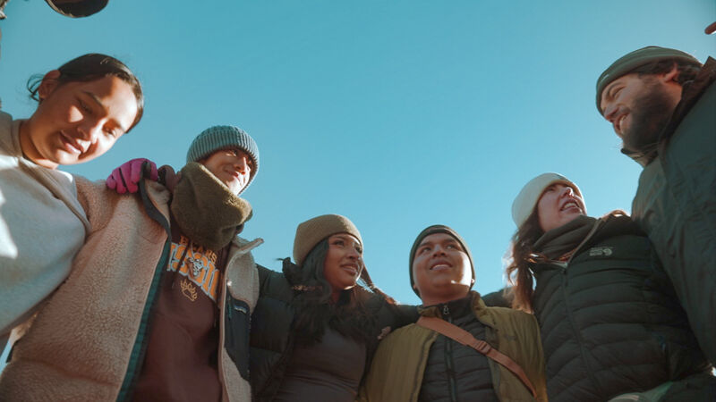 The image shows a group of six people huddled together, looking down at the camera from a low angle. They are dressed in winter clothing, including hats, scarves, and jackets, suggesting a cold environment. The sky is a clear blue, providing a bright backdrop. The composition emphasizes their closeness and unity, capturing a moment of camaraderie and warmth despite the chilly weather.